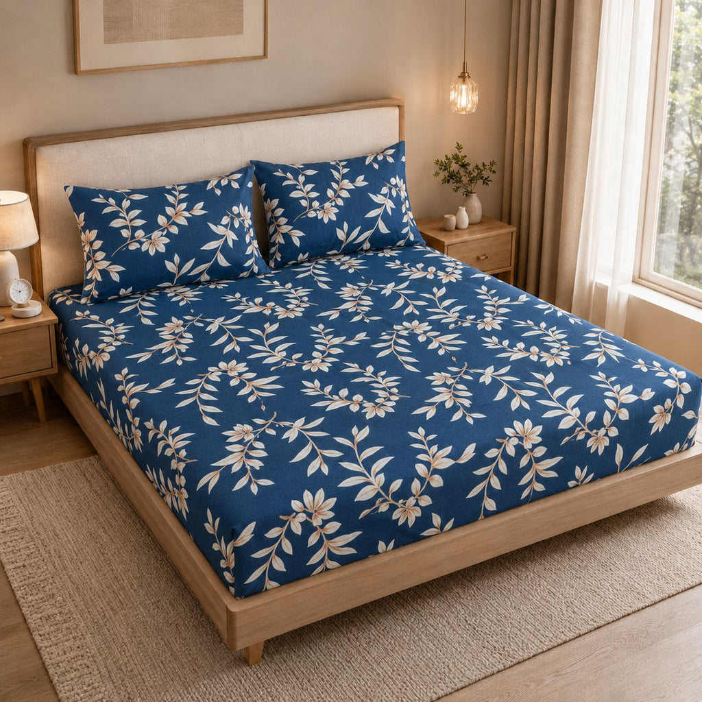 A neatly made bed in a warm, modern bedroom setting, featuring a blue bedding set with a cream floral vine pattern. The bed has a light wood frame and upholstered headboard, flanked by matching bedside tables, a lamp, and a window with beige curtains.