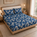A neatly made bed in a warm, modern bedroom setting, featuring a blue bedding set with a cream floral vine pattern. The bed has a light wood frame and upholstered headboard, flanked by matching bedside tables, a lamp, and a window with beige curtains.