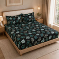 A bed dressed in a black bedding set with a teal and white botanical leaf pattern, styled in a warm, modern bedroom with a wooden headboard, bedside tables, and a window.