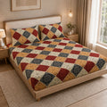 A bed dressed in a patchwork quilted bedspread and matching pillow shams, styled in a warm, modern bedroom setting with wooden furniture, bedside tables, and warm lighting.