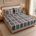 A bed dressed in a patterned sheet set with matching pillowcases, shown in a styled bedroom. The bedding features a geometric patchwork design with gray, dusty pink, and navy blue blocks outlined in white.
