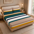 A neatly made bed in a warm-toned bedroom, featuring a striped bedding set with beige, teal, white, and yellow bands. The bed has a light wood frame, matching pillows, and is styled with bedside tables, lamps, and a window with curtains.